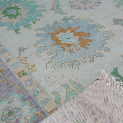 Close-up of a handmade rug with a floral pattern in shades of blue, green, orange, and beige, featuring the brand name 'Paloma' at the bottom.