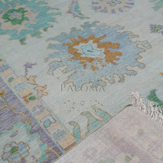 Close-up of a handmade rug with a floral pattern in shades of blue, green, orange, and beige, featuring the brand name 'Paloma' at the bottom.